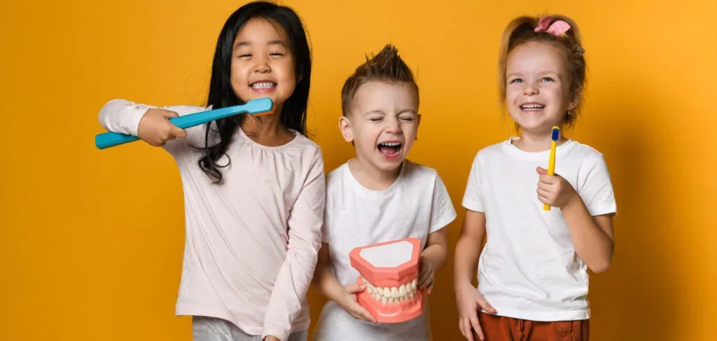 Three children holding toothbrushes and a dental model while learning to brush, building lifelong healthy oral care habits