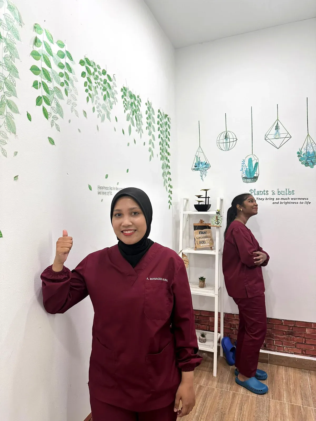 Dental nurse in maroon scrubs beside the plants-and-bulbs feature wall in the Lau & Su Partners Dental clinic, Bukit Jalil