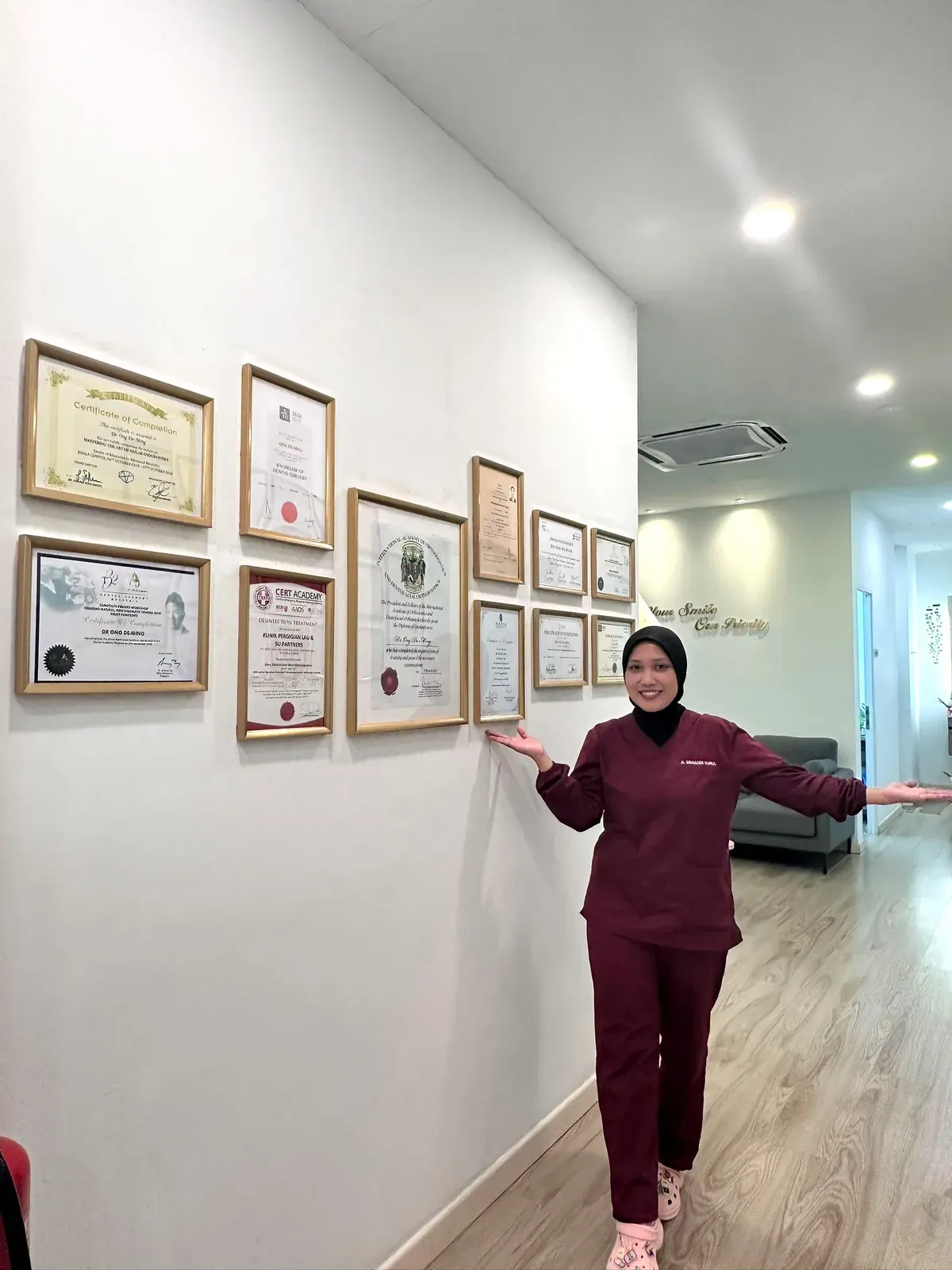 Dental nurse presenting the wall of framed dentist qualifications and certificates in the corridor of our Bukit Jalil clinic
