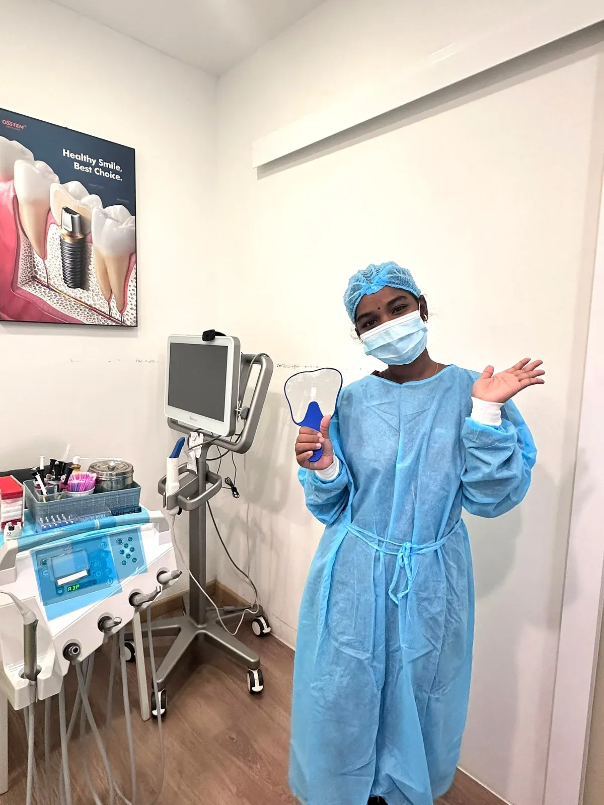 Dental surgical assistant in a sterile blue gown and cap ready beside the instrument trolley in a Bukit Jalil treatment room